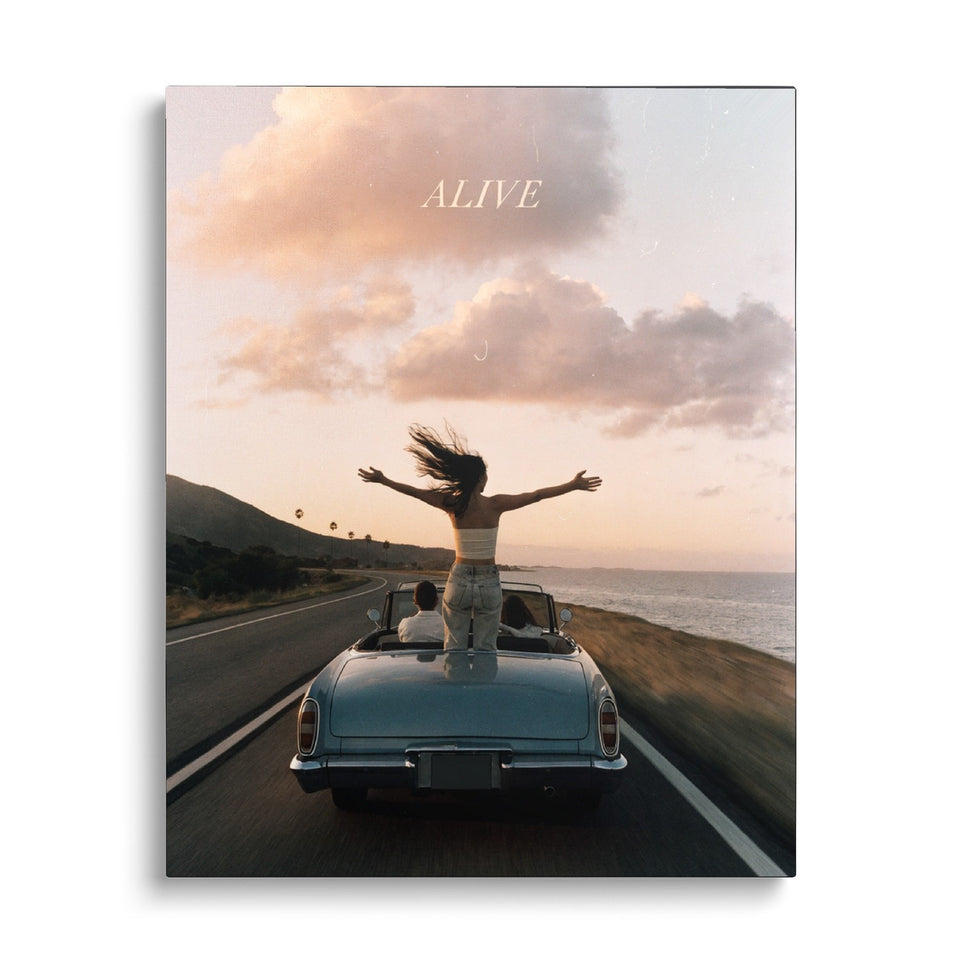 Toile Alive vue de face sur fond blanc : photographie d’une femme debout dans un cabriolet bleu, bras ouverts, sur une route de bord de mer au coucher du soleil