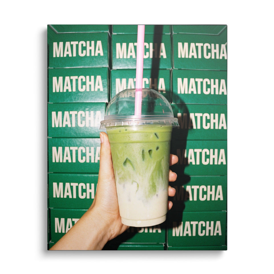 Toile Matcha Latte vue de face sur fond blanc : photographie d’une main tenant un matcha latte glacé à paille rose devant un mur de boîtes vertes marquées « Matcha »