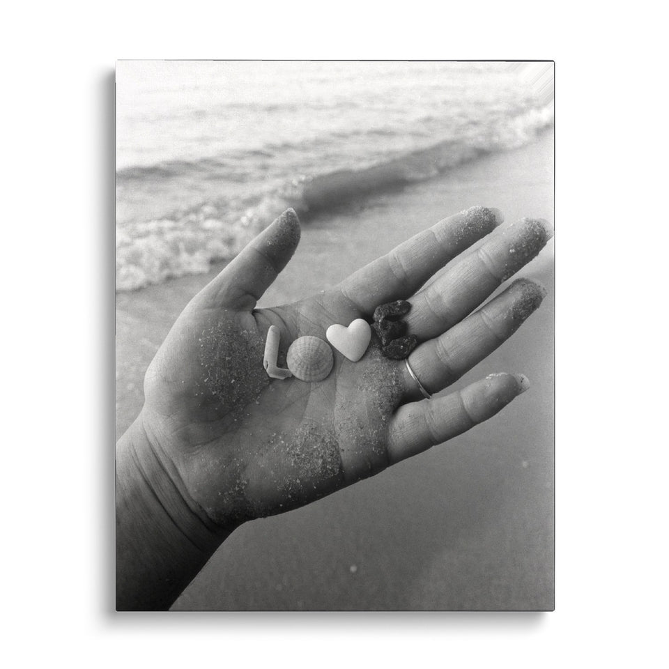 Toile Trésors de Plage vue de face sur fond blanc : photographie noir et blanc d’une main ouverte couverte de sable, tenant des coquillages et galets qui forment le mot « love »