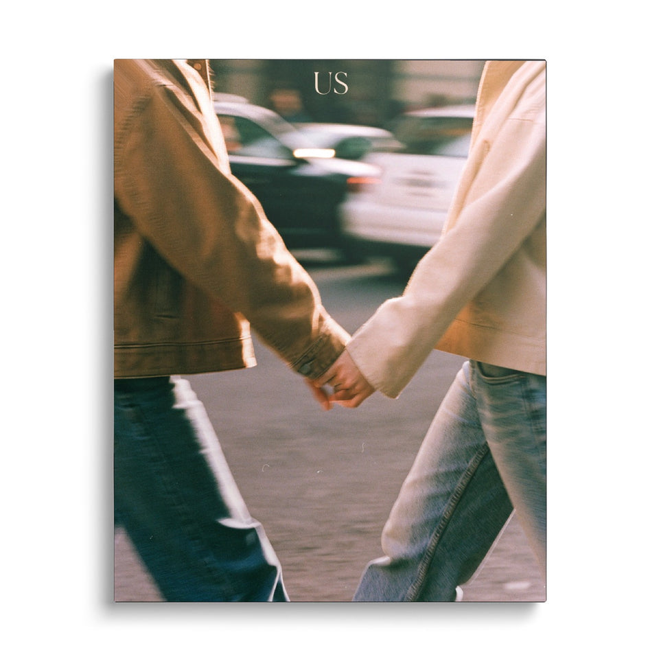 Toile Us vue de face sur fond blanc : photographie de deux personnes en vestes claires traversant une rue main dans la main, voitures floues, mot « Us »