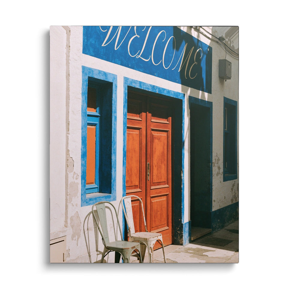 Toile Welcome vue de face sur fond blanc : photographie d’une façade blanche et bleue avec « Welcome » peint à la main, une porte orange et deux chaises en métal